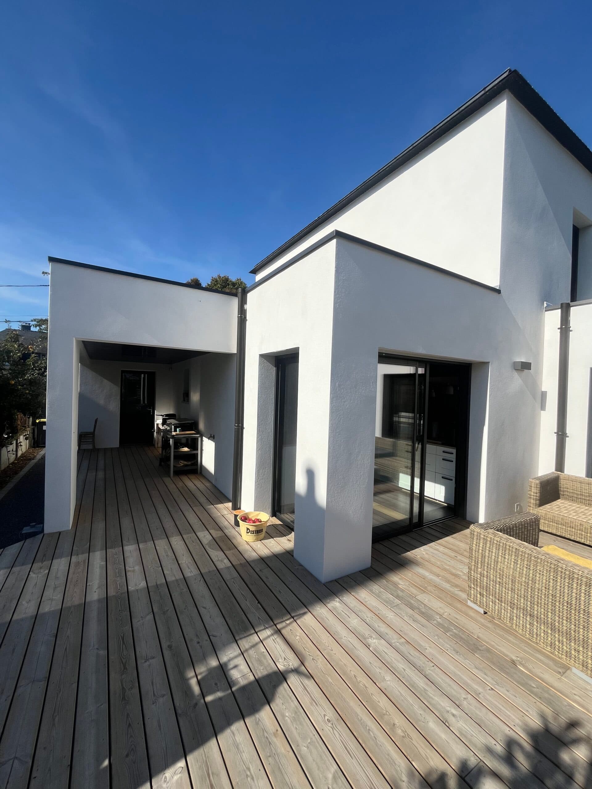 Vue jardin d’une villa moderne avec terrasse bois tout autour, piscine rectangulaire et baies vitrées donnant sur le salon